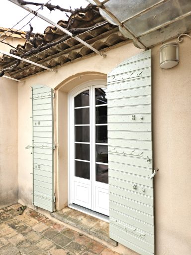Porte blanche avec volets verts, située dans une maison avec un toit en tuiles.