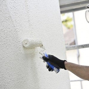 Un homme peint un mur blanc avec un rouleau, portant des gants et des lunettes de protection.