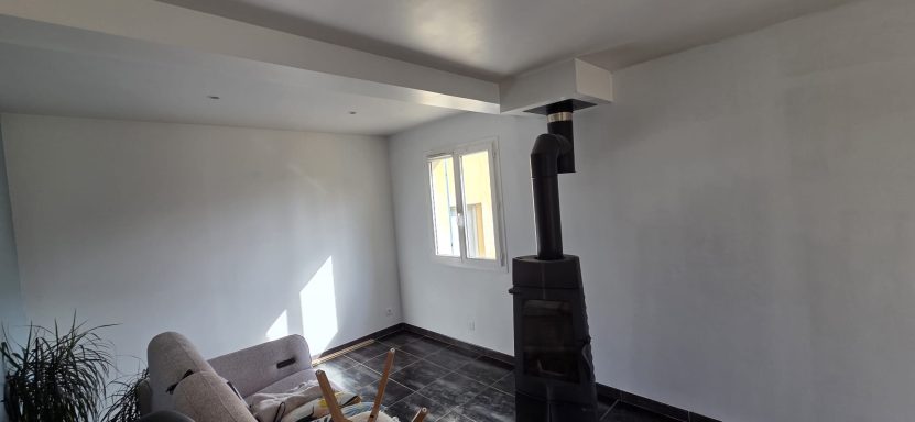 Intérieur d'une pièce avec un poêle, un canapé et une fenêtre laissant entrer la lumière.