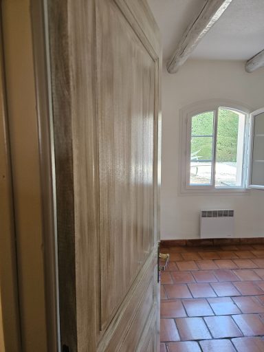 Porte en bois clair, ouverte sur une pièce avec fenêtres et sols carrelés.